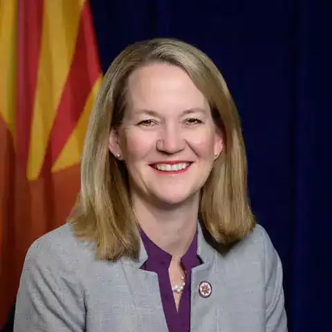 AZ AG Kris Mayes with shoulder-length blonde hair smiles, wearing a gray blazer and purple shirt. Arizona's flag, with its iconic orange and red stripes, is partially visible in the background, hinting at Attorney General Mayes' proud connection to her home state.